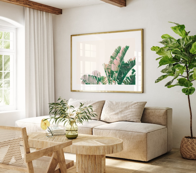 K&ouml;nnte beinhalten: Ein Wohnzimmer-Interieur mit einem beigen Sofa, einem Holz-Couchtisch und einem gerahmten Druck von tropischen Bl&auml;ttern. Der Druck ist in einem goldenen Rahmen und h&auml;ngt an einer wei&szlig;en Wand. In der Ecke des Raumes steht eine gro&szlig;e Topfpflanze.