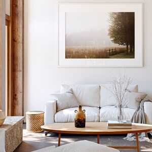 K&ouml;nnte beinhalten: Ein wei&szlig;es Sofa mit zwei Kissen und einer Wolldecke steht in einem Wohnzimmer mit einem Holz-Couchtisch, einem kleinen Beistelltisch und einem gerahmten Landschaftsfoto an der Wand. Eine Vase mit getrockneten Zweigen und eine Keramikvase mit brauner und schwarzer Glasur stehen auf dem Couchtisch.