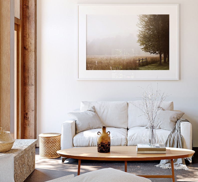 K&ouml;nnte beinhalten: Ein wei&szlig;es Sofa mit zwei Kissen und einer Wolldecke steht in einem Wohnzimmer mit einem Holz-Couchtisch, einem kleinen Beistelltisch und einem gerahmten Landschaftsfoto an der Wand. Eine Vase mit getrockneten Zweigen und eine Keramikvase mit brauner und schwarzer Glasur stehen auf dem Couchtisch.