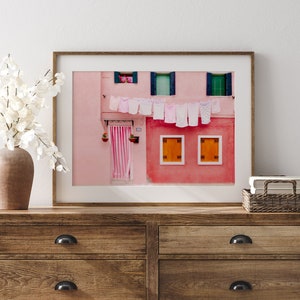 Peut inclure: Une photographie encadr&eacute;e d'un b&acirc;timent rose avec du linge blanc suspendu &agrave; une corde &agrave; linge. Le b&acirc;timent a des fen&ecirc;tres vertes et des volets orange.