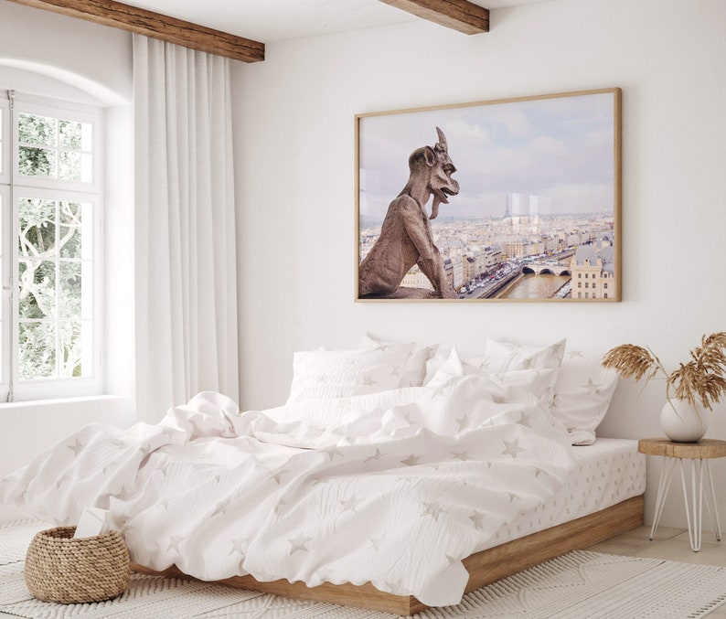 May include: A white bed with a white duvet cover and white pillowcases. The duvet cover has a subtle star pattern. The bed is in a bedroom with a window and a picture on the wall. The picture shows a gargoyle overlooking a city.