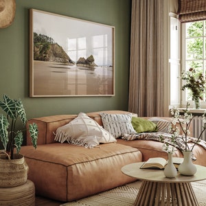 May include: A light brown leather sectional sofa with a variety of throw pillows, including a white pillow with fringe, a black and white patterned pillow, and a green pillow. The sofa is in a living room with a framed photograph of a beach scene, a window with white blinds, and a wooden coffee table with a book and two vases.