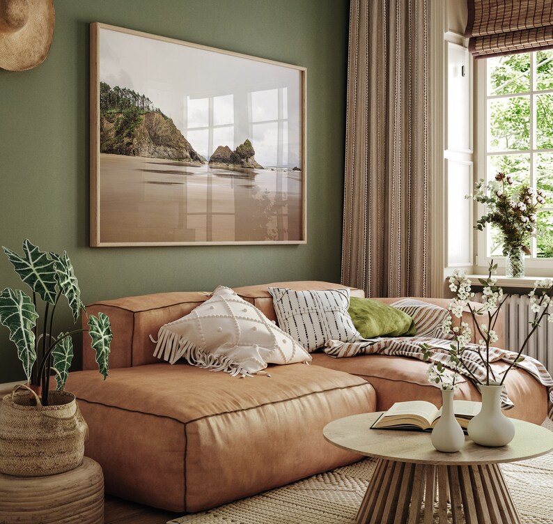 May include: A light brown leather sectional sofa with a variety of throw pillows, including a white pillow with fringe, a black and white patterned pillow, and a green pillow. The sofa is in a living room with a framed photograph of a beach scene, a window with white blinds, and a wooden coffee table with a book and two vases.