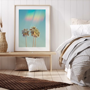 May include: Framed art print featuring three palm trees against a light blue sky with a rainbow effect. The artwork is displayed on a light wood bench with pillows and a woven vase. The room has a neutral color palette with a bed and a brown rug.