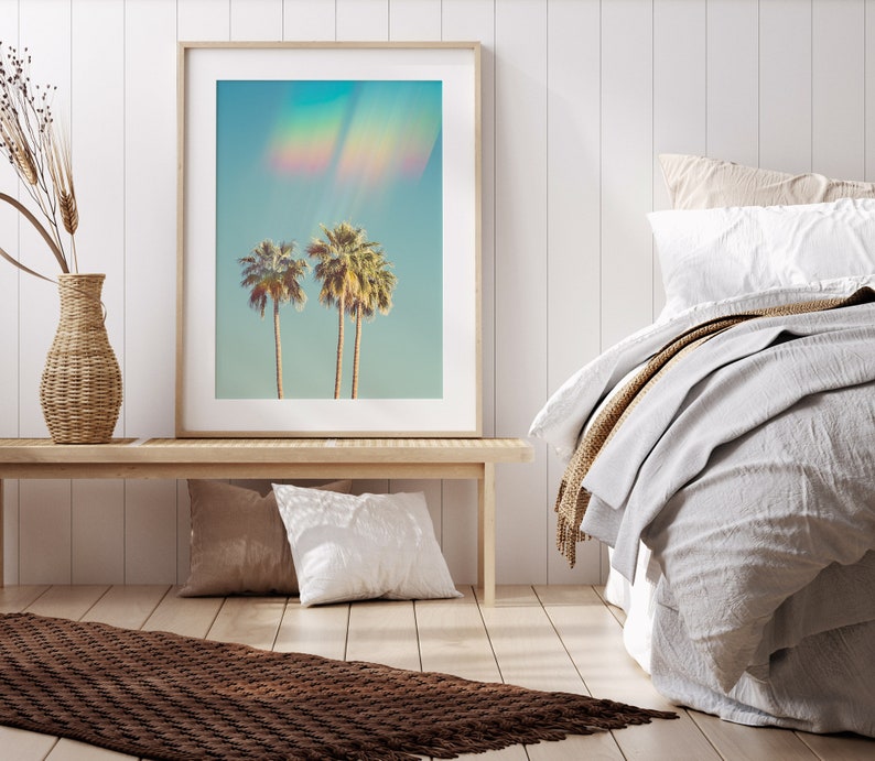 May include: Framed art print featuring three palm trees against a light blue sky with a rainbow effect. The artwork is displayed on a light wood bench with pillows and a woven vase. The room has a neutral color palette with a bed and a brown rug.