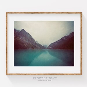 May include: A framed photograph of a mountain lake with two mountain peaks in the background. The water is a vibrant teal color and the sky is a soft gray. The mountains are a dark gray color and the image has a vintage feel.