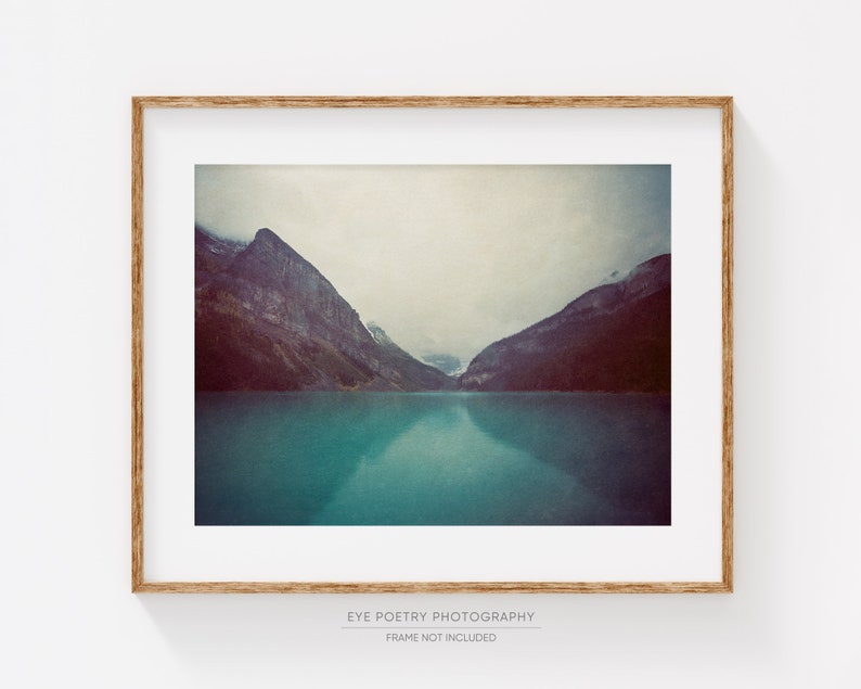 May include: A framed photograph of a mountain lake with two mountain peaks in the background. The water is a vibrant teal color and the sky is a soft gray. The mountains are a dark gray color and the image has a vintage feel.