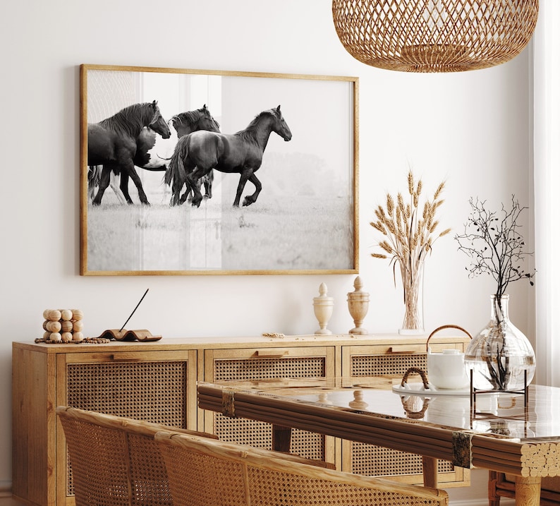 May include: Black and white photograph of four horses running in a field. The photo is framed and hanging on a wall above a wooden cabinet with woven cane doors.