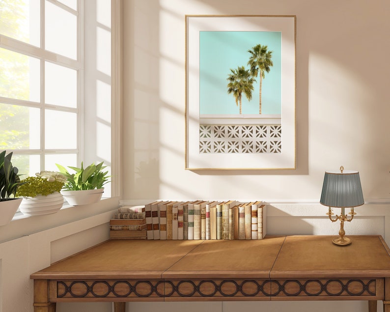 May include: A wooden desk with a decorative border, a framed print of palm trees against a blue sky, and a gold table lamp with a gray pleated shade.