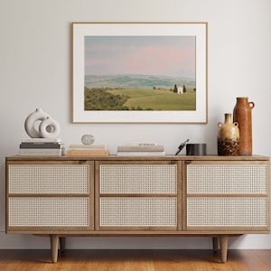 May include: A wooden cabinet with three drawers, each with a woven rattan front. The cabinet is decorated with a framed photograph of a white building in a green field, a ceramic sculpture, and two ceramic vases.