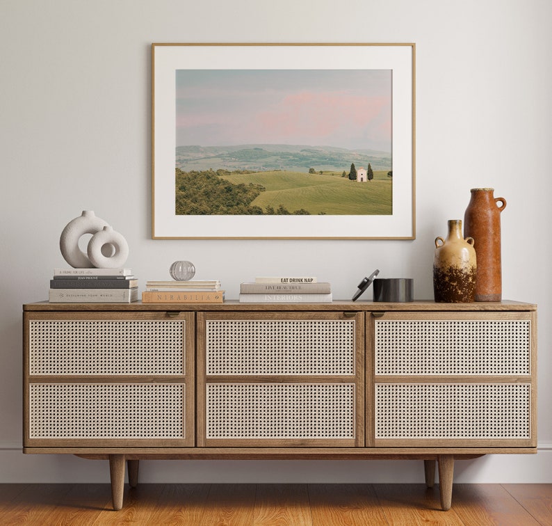 May include: A wooden cabinet with three drawers, each with a woven rattan front. The cabinet is decorated with a framed photograph of a white building in a green field, a ceramic sculpture, and two ceramic vases.