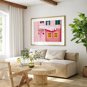 Peut inclure: Un int&eacute;rieur de salon avec un canap&eacute; beige en forme de L, une table basse en bois et une impression encadr&eacute;e d'un b&acirc;timent rose avec du linge suspendu &agrave; une ligne. Une plante en pot avec des feuilles vertes se trouve dans le coin de la pi&egrave;ce.
