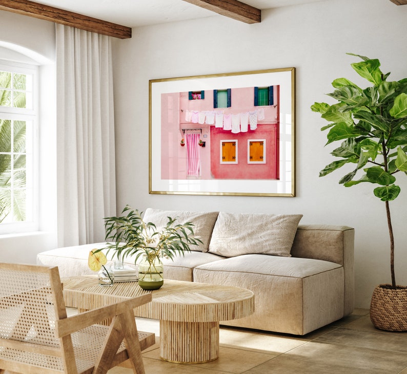 Peut inclure: Un int&eacute;rieur de salon avec un canap&eacute; beige en forme de L, une table basse en bois et une impression encadr&eacute;e d'un b&acirc;timent rose avec du linge suspendu &agrave; une ligne. Une plante en pot avec des feuilles vertes se trouve dans le coin de la pi&egrave;ce.