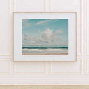 May include: A framed photograph of a beach scene with a large white cloud in the sky and blue water with waves crashing on the shore.