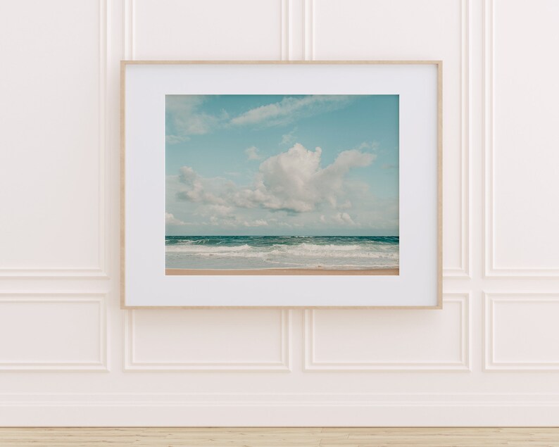 May include: A framed photograph of a beach scene with a large white cloud in the sky and blue water with waves crashing on the shore.