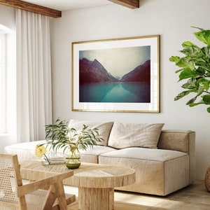 May include: A living room interior with a light beige sectional sofa, a wooden coffee table, and a framed photograph of a mountain lake. A large potted plant with green leaves is in the corner of the room.