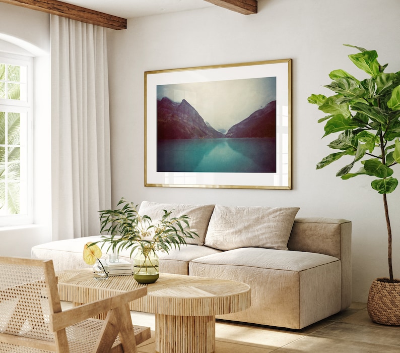 May include: A living room interior with a light beige sectional sofa, a wooden coffee table, and a framed photograph of a mountain lake. A large potted plant with green leaves is in the corner of the room.