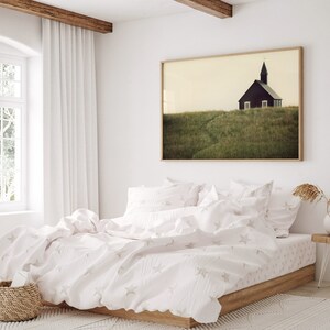 K&ouml;nnte beinhalten: Ein wei&szlig;es Bett mit einer wei&szlig;en und grauen Bettw&auml;sche mit Sternenmuster und Kissen. Das Bett steht in einem Schlafzimmer mit einem gro&szlig;en Fenster und einem gerahmten Druck einer kleinen Kirche auf einem grasbewachsenen H&uuml;gel.