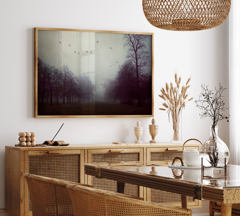 May include: A framed landscape art print depicting a misty forest scene with trees and flying birds. The artwork is displayed above a wooden cabinet with woven rattan doors. Decorative items include vases and a woven pendant light.