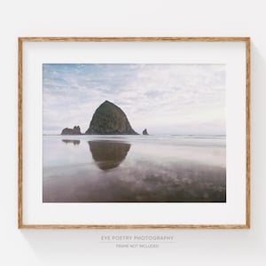 May include: A photograph of a large rock formation jutting out of the ocean, with a reflection of the rock in the water. The sky is a soft blue with white clouds.