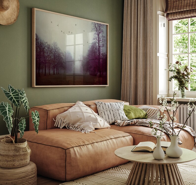 May include: A cozy living room scene featuring a large, framed landscape art piece with trees in a misty setting. A brown leather sectional sofa is adorned with decorative pillows. A round wooden coffee table and potted plants complete the inviting space.