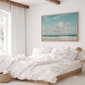 May include: A white bed with a white duvet cover and white pillowcases featuring a subtle star pattern. The bed is made up with a white throw blanket and has a wooden frame. A framed photograph of a beach scene hangs on the wall above the bed.