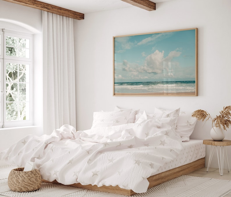May include: A white bed with a white duvet cover and white pillowcases featuring a subtle star pattern. The bed is made up with a white throw blanket and has a wooden frame. A framed photograph of a beach scene hangs on the wall above the bed.