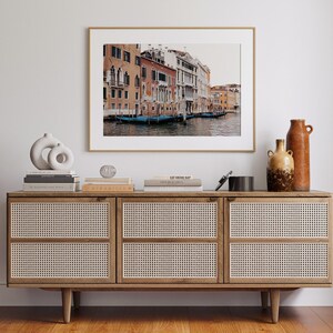 May include: A framed photograph of a canal in Venice, Italy. The photo shows a row of colorful buildings along the canal with gondolas docked in the water. The photo is hanging on a white wall above a wooden dresser with woven rattan doors.