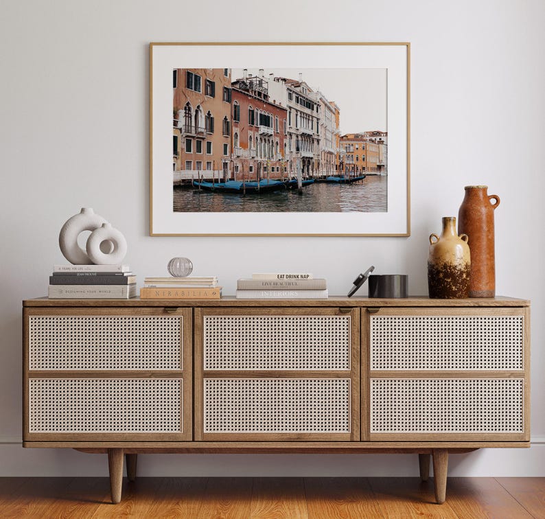 May include: A framed photograph of a canal in Venice, Italy. The photo shows a row of colorful buildings along the canal with gondolas docked in the water. The photo is hanging on a white wall above a wooden dresser with woven rattan doors.