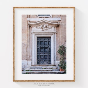 May include: A vintage-style photograph of a dark wooden door with a carved stone frame. The door is set in a building with a light-colored facade. The door is surrounded by steps and a small bush.