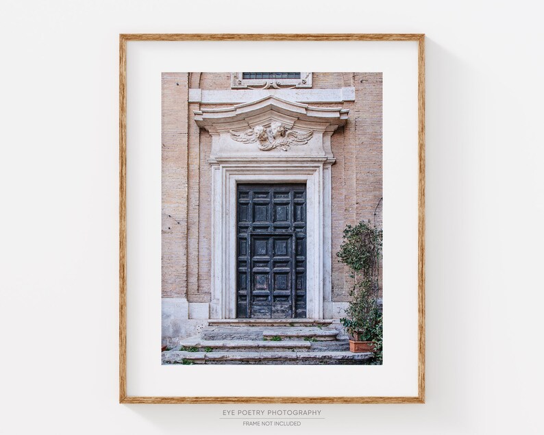 May include: A vintage-style photograph of a dark wooden door with a carved stone frame. The door is set in a building with a light-colored facade. The door is surrounded by steps and a small bush.