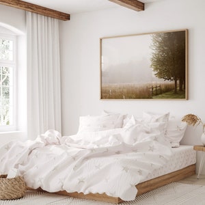 K&ouml;nnte beinhalten: Ein wei&szlig;es Bett mit einem wei&szlig;en und grauen Sternenmuster-Bettbezug und passenden Kissenbez&uuml;gen. Das Bett ist mit einem wei&szlig;en Laken und einer wei&szlig;en Decke bezogen. Das Bett steht in einem Schlafzimmer mit einem gro&szlig;en Fenster und einem gerahmten Bild eines nebligen Feldes mit einem Baum an der Wand.