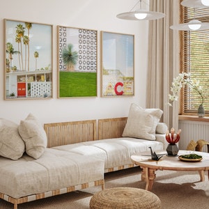 May include: A white couch with three pillows sits in front of a white wall with three framed prints. The prints are of a palm tree landscape, a geometric pattern, and a cityscape. A wooden coffee table with a black origami crane sits in front of the couch.