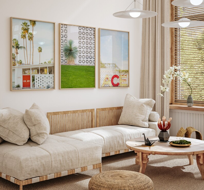 May include: A white couch with three pillows sits in front of a white wall with three framed prints. The prints are of a palm tree landscape, a geometric pattern, and a cityscape. A wooden coffee table with a black origami crane sits in front of the couch.