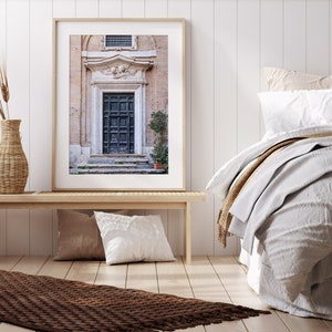 May include: A framed photograph of a vintage wooden door with a carved stone archway above it. The door is dark brown and has a weathered look. The archway is decorated with intricate carvings. The photo is set against a white wall and a bed with white linens.