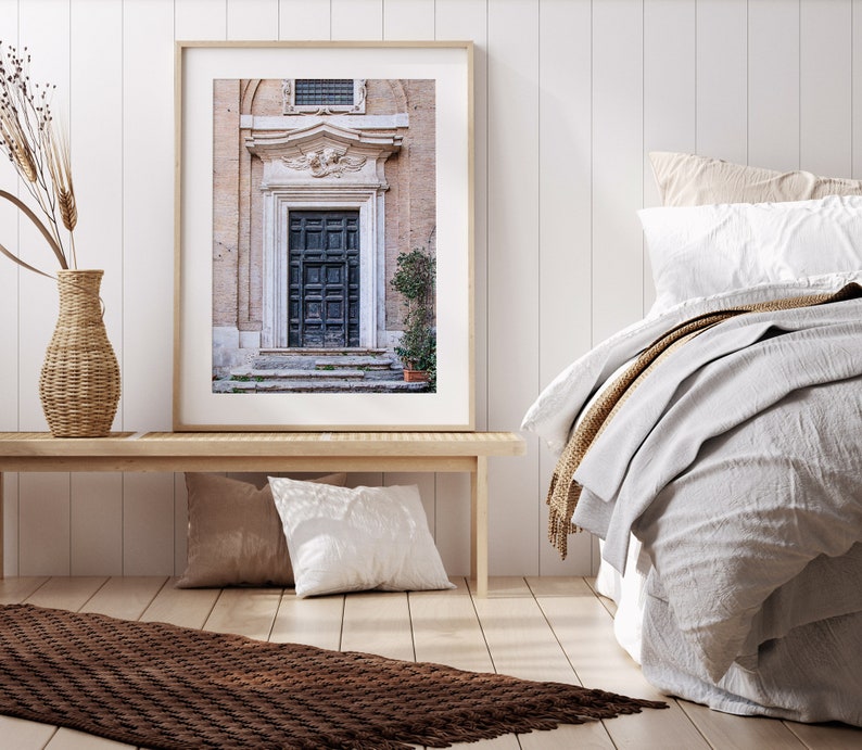 May include: A framed photograph of a vintage wooden door with a carved stone archway above it. The door is dark brown and has a weathered look. The archway is decorated with intricate carvings. The photo is set against a white wall and a bed with white linens.