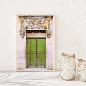 May include: A framed photograph of a green wooden door with ornate carvings above it. The door is set in a pink building with peeling paint.