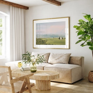 May include: A living room interior with a beige sectional sofa, a wooden coffee table, and a framed landscape photograph of rolling hills and a small building. A potted plant with green leaves is in the corner of the room.