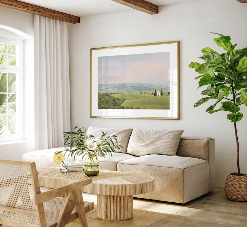May include: A living room interior with a beige sectional sofa, a wooden coffee table, and a framed landscape photograph of rolling hills and a small building. A potted plant with green leaves is in the corner of the room.