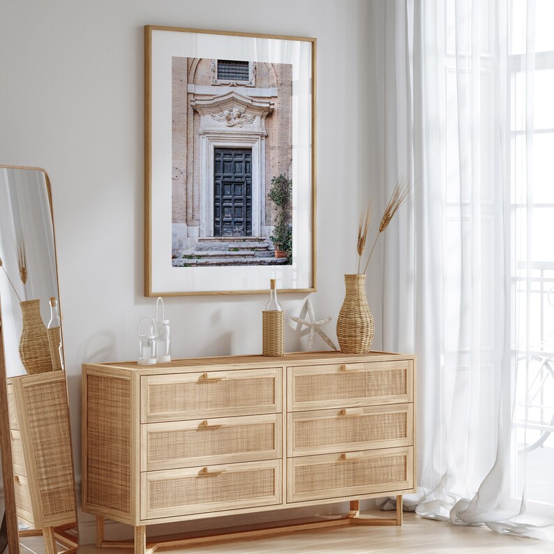 May include: A light wood dresser with six drawers, a framed photograph of an old doorway, and a vase with dried flowers on top. The dresser is in a room with white walls and a white sheer curtain.
