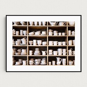 May include: A white wooden shelf unit filled with various white ceramic mugs, cups, bowls, and pitchers. The shelf unit is divided into 18 square compartments.