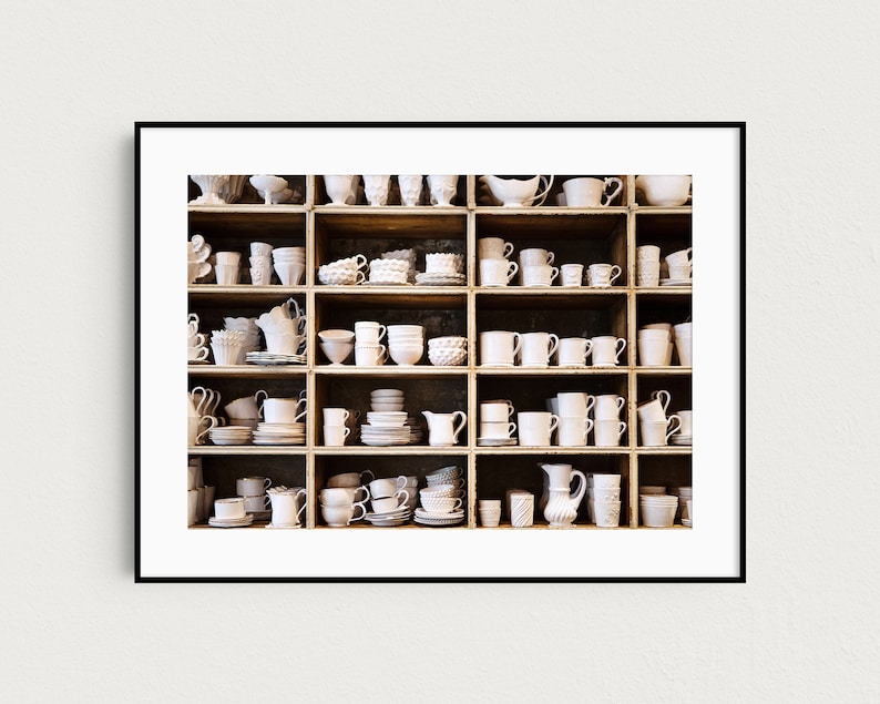 May include: A white wooden shelf unit filled with various white ceramic mugs, cups, bowls, and pitchers. The shelf unit is divided into 18 square compartments.