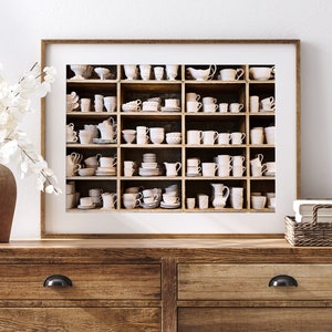 May include: A framed print of a white shelf filled with various white ceramic cups, saucers, and bowls. The shelf is divided into nine sections, each with a different arrangement of items. The print is set against a white wall and a wooden dresser.
