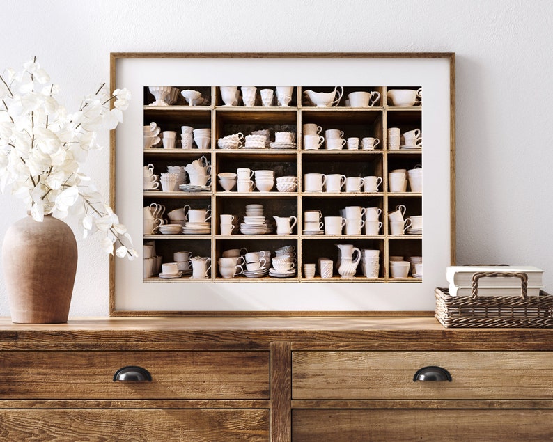 May include: A framed print of a white shelf filled with various white ceramic cups, saucers, and bowls. The shelf is divided into nine sections, each with a different arrangement of items. The print is set against a white wall and a wooden dresser.