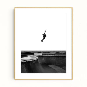 Peut inclure: Photographie en noir et blanc d'un skateboarder en l'air au-dessus d'un bowl de skatepark. L'image est encadr&eacute;e dans un cadre dor&eacute;, avec un fond blanc. Le skatepark pr&eacute;sente des structures en b&eacute;ton incurv&eacute;es.