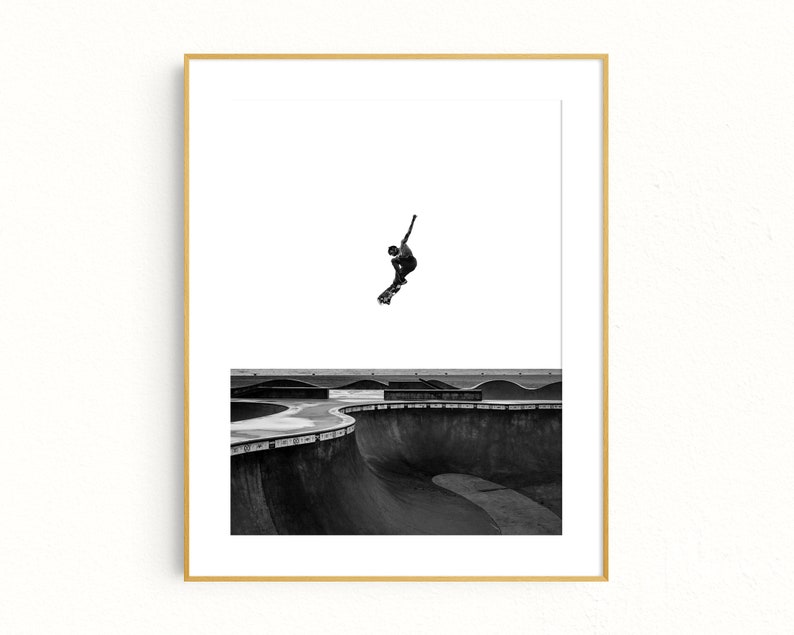 Peut inclure: Photographie en noir et blanc d'un skateboarder en l'air au-dessus d'un bowl de skatepark. L'image est encadr&eacute;e dans un cadre dor&eacute;, avec un fond blanc. Le skatepark pr&eacute;sente des structures en b&eacute;ton incurv&eacute;es.