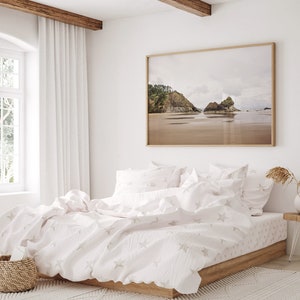 May include: A white bed with a white and gray star patterned duvet cover and matching pillowcases. The bed is made with a wooden frame and is sitting on a white rug. A large framed photograph of a beach scene hangs on the wall behind the bed.