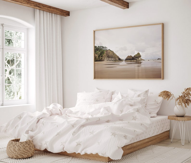May include: A white bed with a white and gray star patterned duvet cover and matching pillowcases. The bed is made with a wooden frame and is sitting on a white rug. A large framed photograph of a beach scene hangs on the wall behind the bed.