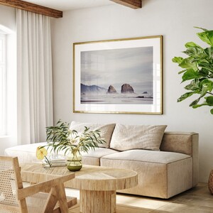 May include: A living room interior with a beige sectional sofa, a wooden coffee table, a wicker chair, and a large framed photograph of a beach scene with rock formations. A potted plant with large green leaves is in the corner of the room.