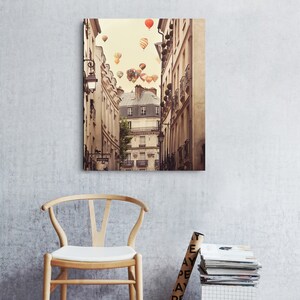 May include: A photograph of a narrow street in Paris, France, with colorful hot air balloons floating above the buildings. The buildings are old and have a vintage look. The street is lined with trees and there are people walking on the sidewalk.